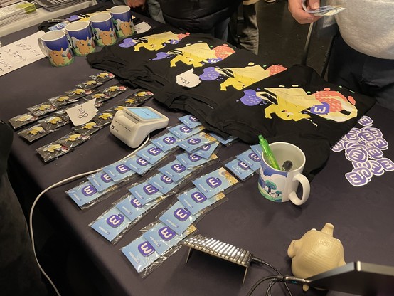 Our desk full of t-shirts, pins, mugs, and stickers, with people queuing up behind it.
